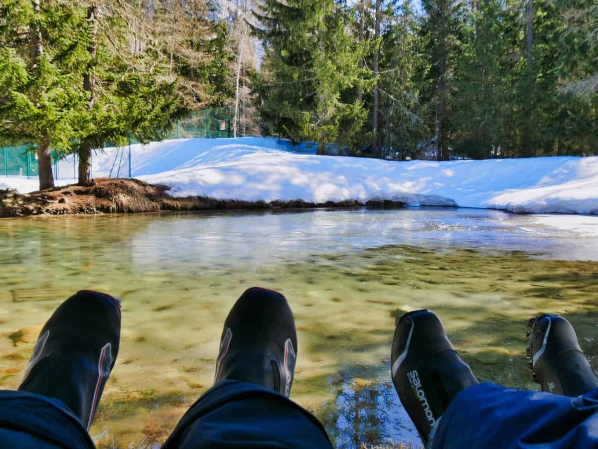 Two sets of feet up looking out over clear lake with snow and trees surrounding it