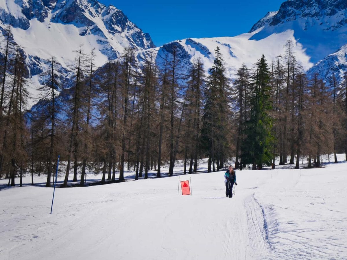 Kalyn walking carrying her Nordic Skis surrounded by snowy mountains and trees in the Nordic Ski Area in The French Alps