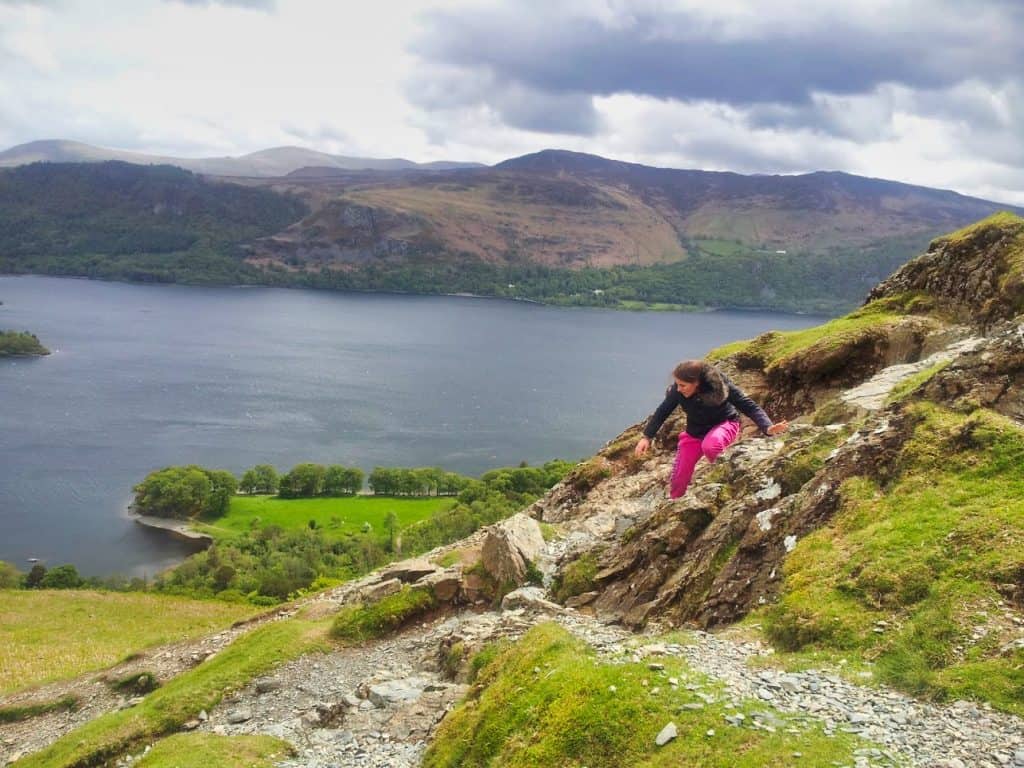 Kalyn climbing down mountain in Lake District, wearing pink pants and a blue coat with a lake behind her. 