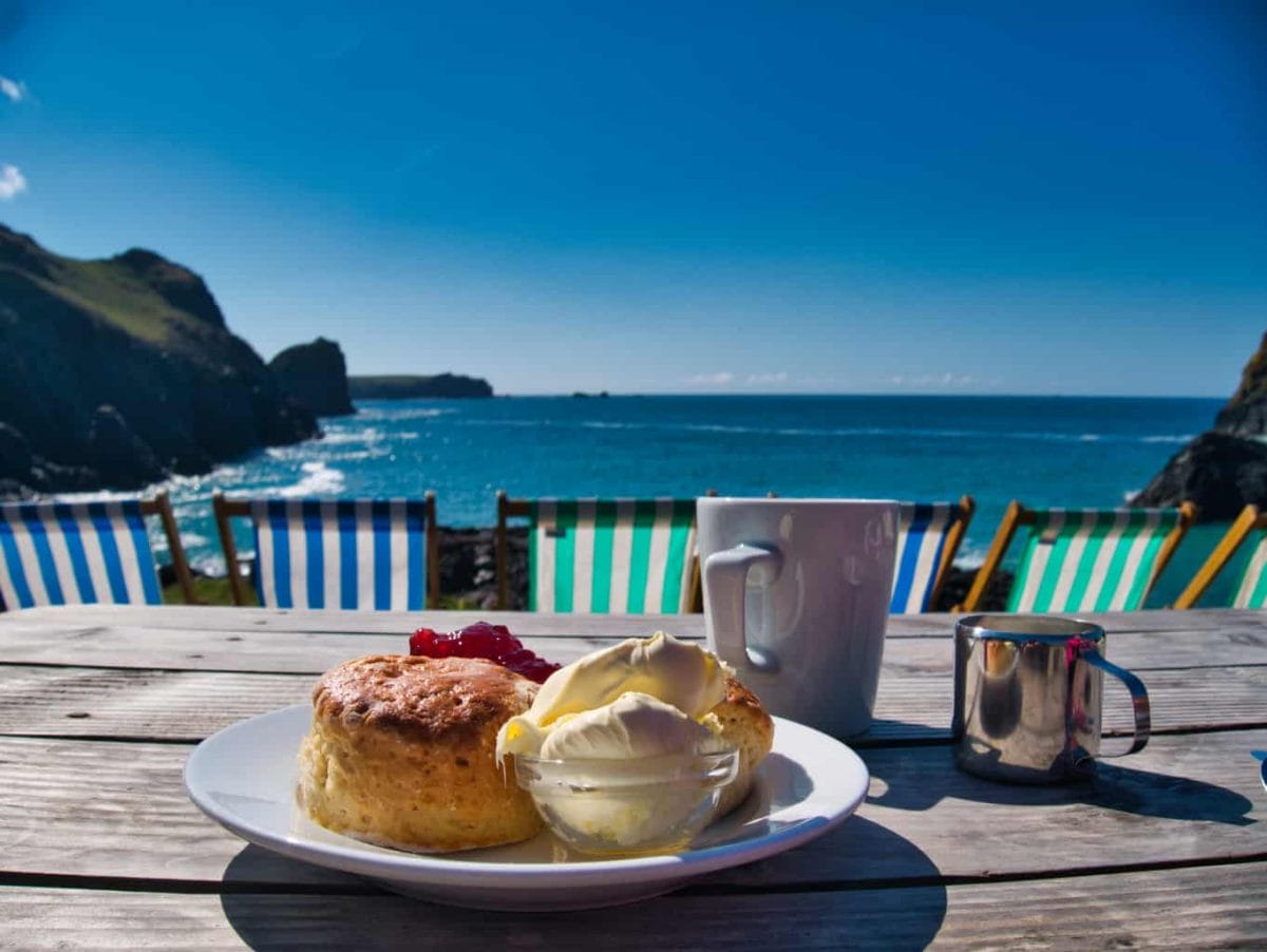 A cream tea with a scone jam and clotted cream and cup of tea with blue sky in the background deep blue sea and some deck chairs
