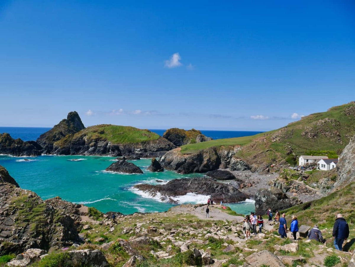 Deep blue water with rocks sticking out and the cafe at Kynance Cove with people walking up a path