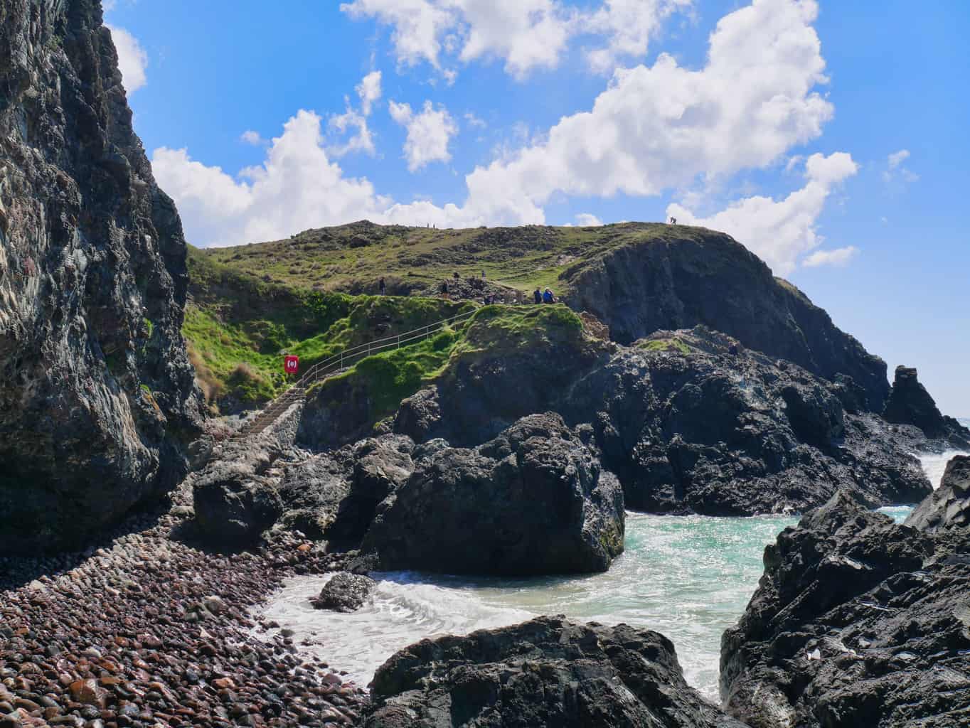 A rocky path towards the cafe at Kynance Cove with water coming up to the cliff and green hills in the background