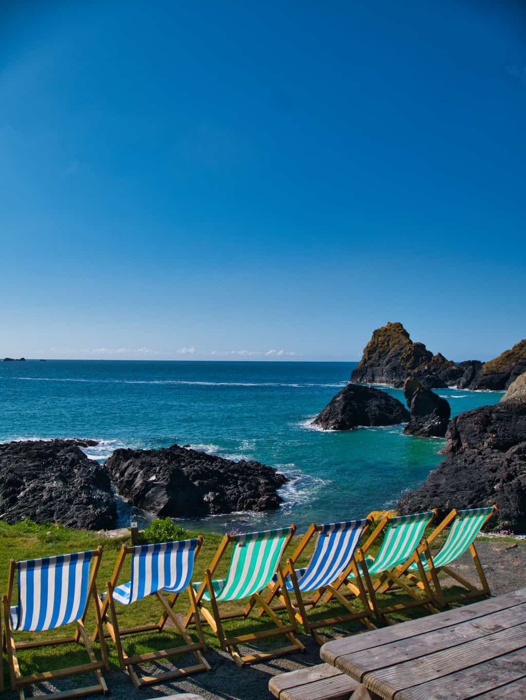 Blue and green deck chairs looking out over the blue water at Kynance Cove with blue sky