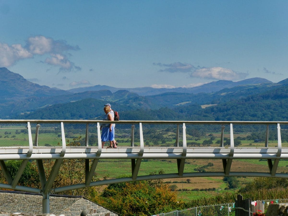 The bridge that crosses over to Harlech Castle with two people walking on it and the Snowdonia mountains in the distance