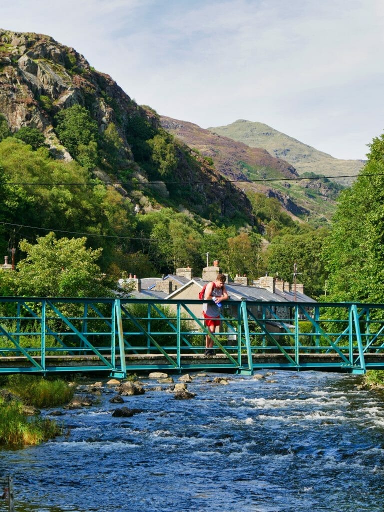 A man on bridge in Beddgelert Wales with a river flowing underneath and hills in the background