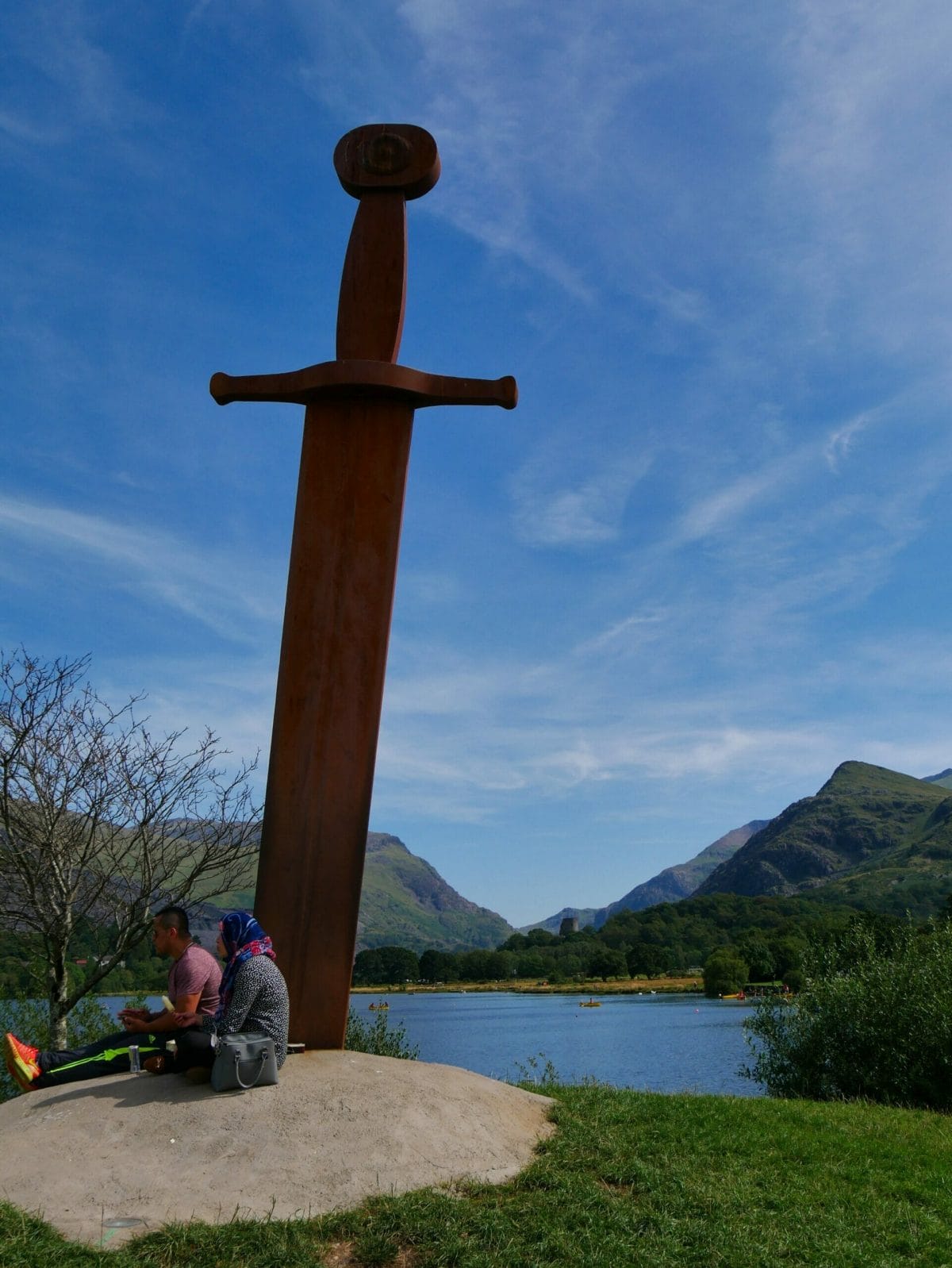 Sword monument sticking out of the ground with people leaning against it in Wales with lake behind it