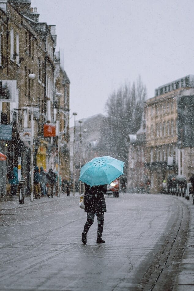 Person crossing the road in London as it snows, holding a blue umbrella