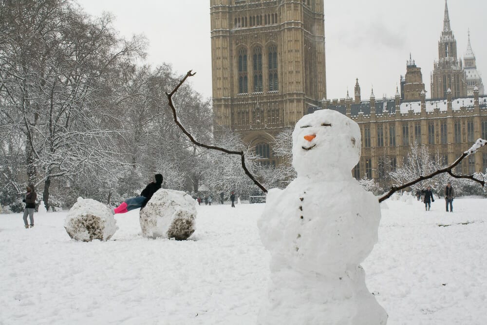 A snowman with a smile in front of Parliament, with its arms raised, with people playing in the snow behind