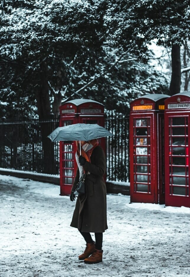 Woman with umbrella walking on snow in London in front of red phone booths