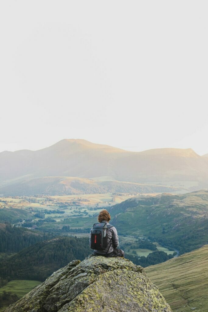 Man with brown hair and a backpack on sitting on a rock in the Lake District, looking out to the Lake District in front of him as we see him from behind.